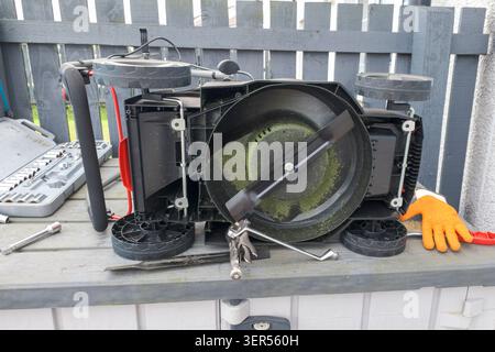 Broken Lawn mower rests on a workbench outdoors. Tools and gloves nearby suggest repair or disassembly. Fence behind adds urban or backyard setting. I Stock Photo
