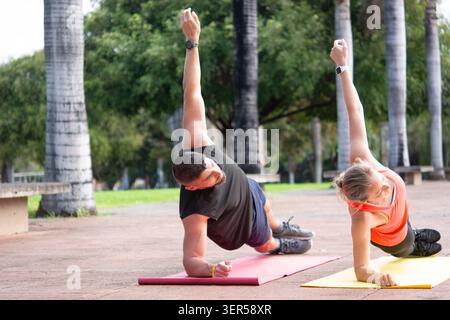 Mature man and woman practicing side plank exercises on yoga mats in a sunny park. Stock Photo