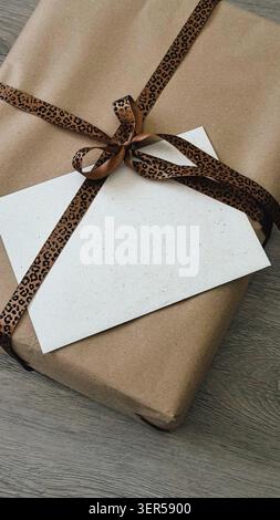 Brown gift package wrapped in kraft paper with a leopard print ribbon and a blank card placed on a wooden Stock Photo