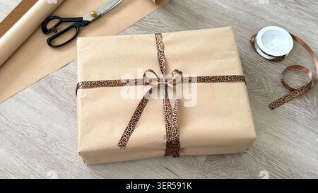 Wrapped gift box with leopard print ribbon and scissors on wooden surface, additional wrapping paper and tape visible in the background Stock Photo