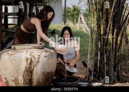 Two happy women cleaning dishes together using water from a large traditional clay jar in a rural outdoor setting showcasing sustainable living. Stock Photo