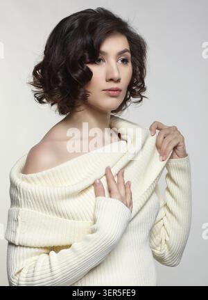 Brunette beautiful woman dressed in large white woolen sweater on gray background. Female with hands on sweater collar Stock Photo