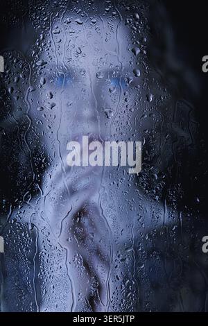 Sad young woman behind the window glass with raindrops. Female in retro vintage dress looking through the glass on black background. Focus on water dr Stock Photo