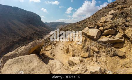 Rocky mountain trail on a dry desert hillside with rugged cliffs, loose stones and blue sky with clouds, showing an arid landscape and remote hiking r Stock Photo