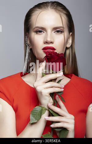 Female with red rose in hand and long earring with rhinestones in the ear. Portrait of young woman in red dress and a strand of hair on the face Stock Photo
