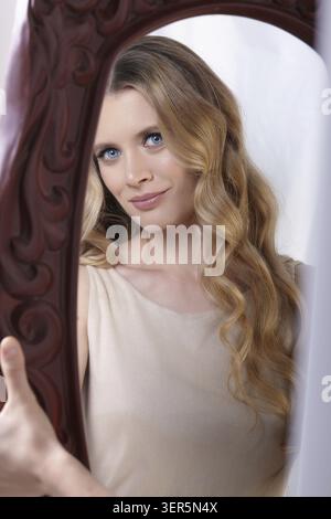 Female holds in her hands a large mirror in a brown frame. A woman looks in the mirror. The woman is wearing a cream beige dress with straps. Female b Stock Photo