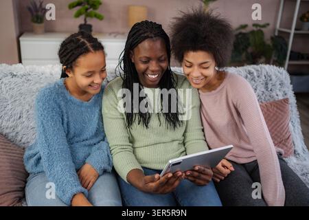 Three excited girls gather around their mother, sharing smiles and laughter as they watch a fun video on a tablet in a warm, inviting living room fill Stock Photo