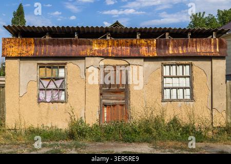 Weathered building features yellow stucco facade and rusty corrugated metal roof. Overgrown weeds and dry grass enhance the rustic, neglected atmosphe Stock Photo