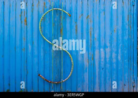 Large green dollar sign sculpture mounted on bright blue corrugated metal. Rust and wear on sculpture highlight age against textured industrial backgr Stock Photo