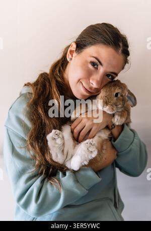 Young woman lovingly holding a fluffy brown dwarf rabbit against a simple background Stock Photo