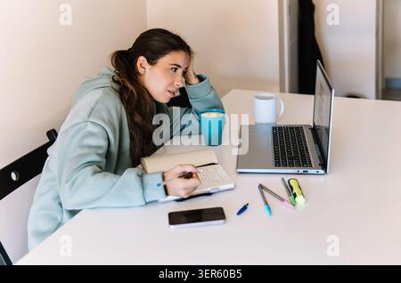 Young woman working from home, concentrating while taking notes and using a laptop Stock Photo