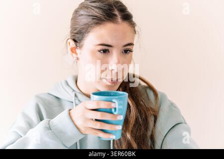 Woman in a hoodie drinking from a blue mug, looking thoughtful Stock Photo