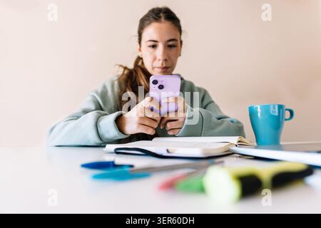 A woman is sitting at a table with a cell phone in her hand. She is looking at the phone and she is focused on it. The table is cluttered with various Stock Photo