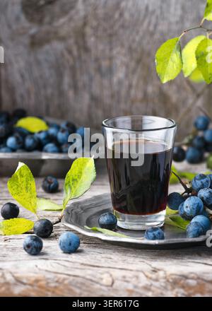 Homemade healthy sloe berry beverage in glass on rustic wooden background. Dark blue fresh harvested blackthorn fruits (Prunus spinosa) Stock Photo
