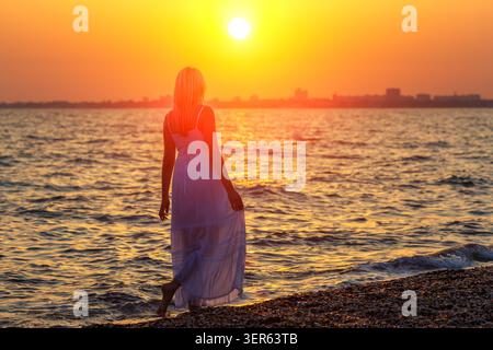 Young blonde woman in a flowing white dress walking barefoot along the shoreline silhouetted against a vivid orange sunset sky over the sea at dusk Stock Photo