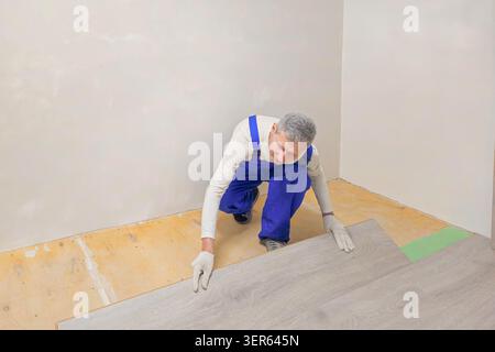 A man in a blue jumpsuit is laying down a piece of wood laminate flooring. The man is wearing gloves and a blue jumpsuit Stock Photo