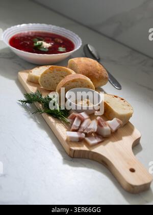 Bowl of traditional borscht soup topped with sour cream and herbs. Sliced fresh bread, crispy bacon pieces and dill garnish on wooden cutting Stock Photo