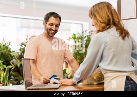 Smiling florist holding credit card reader while female customer paying with smartphone using nfc technology Stock Photo