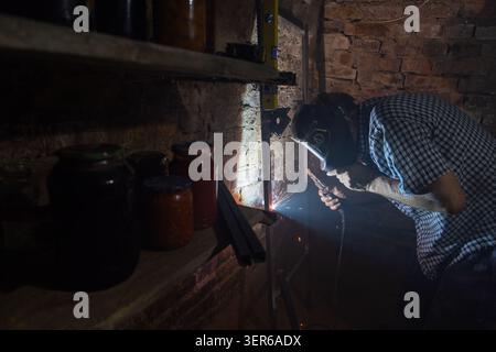 Man actively welds metal in dimly lit workshop. Sparks illuminate the scene, highlighting shelves of glass jars and a brick wall. Stock Photo