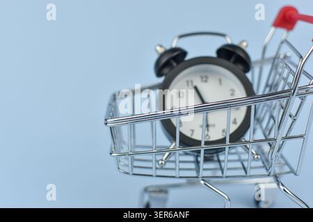 Mini clock sits in shopping cart. Metal cart holds timepiece with black frame. White dial, black hands, simple numerals. Soft blue background emphasiz Stock Photo