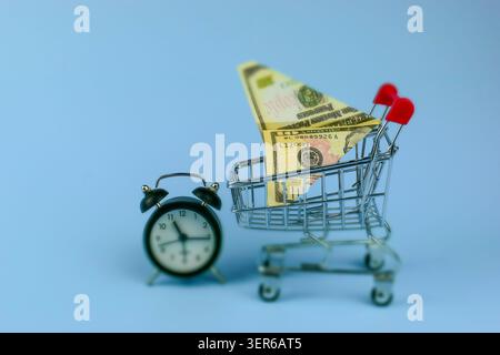 Mini shopping cart holds folded cash bills. Alarm clock rests beside it on plain blue background. Scene implies time-sensitive spending or budgeting. Stock Photo