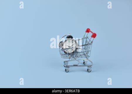 Mini clock sits in shopping cart. Metal cart holds timepiece with black frame. White dial, black hands, simple numerals. Soft blue background emphasiz Stock Photo