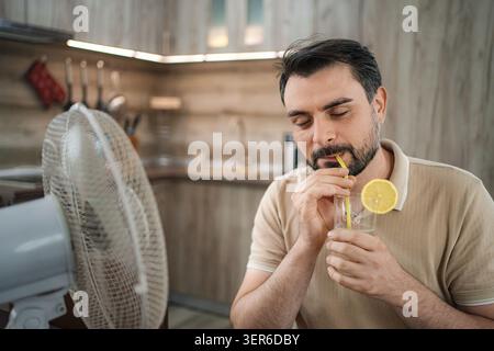 Man refreshing with a cold drink in front of a fan during a summer heat wave at home Stock Photo
