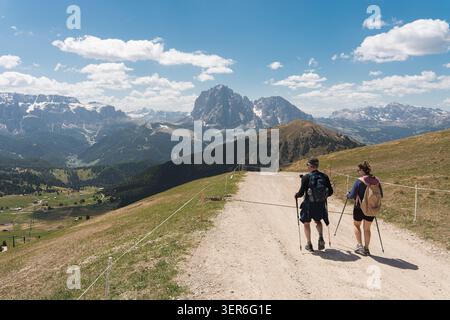 Man and woman hiking with backpacks and trekking poles along a wide dirt road on a high mountain ridge overlooking a vast alpine valley, outdoor explo Stock Photo