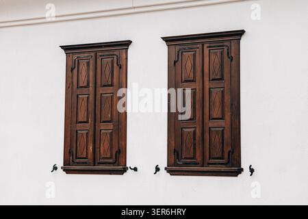 Two closed vintage wooden window shutters with decorative iron hardware on a minimal white wall facade, showing old architecture details Stock Photo
