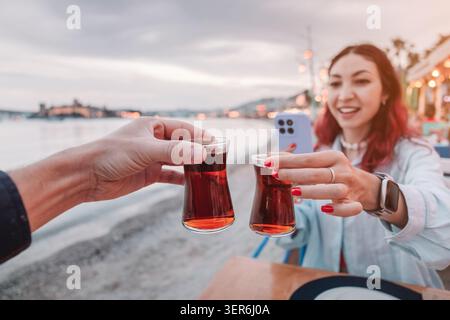 Young couple clinking traditional Turkish tea glasses outdoors at sunset, enjoying vacation lifestyle with Bodrum Castle in background Stock Photo