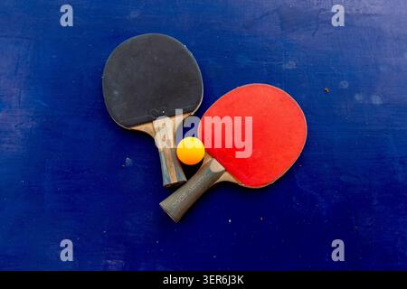 Two table tennis rackets—one red and one black—resting next to a bright orange ping pong ball on a blue playing surface. The worn outdoor table featur Stock Photo