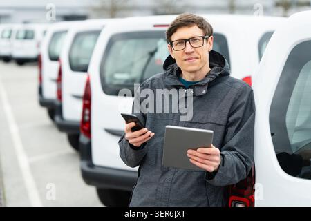 Fleet manager with digital tablet and smartphone supervising rows of white delivery vans. Stock Photo