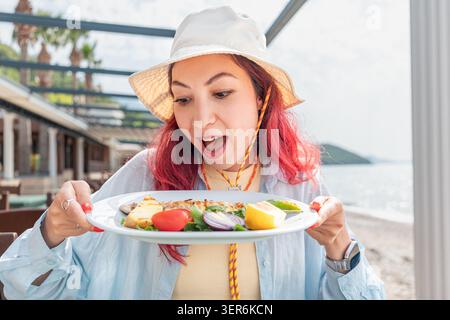 Young woman with red hair wearing a bucket hat, looking down enthusiastically at a plate of grilled fish at a beachfront restaurant Stock Photo