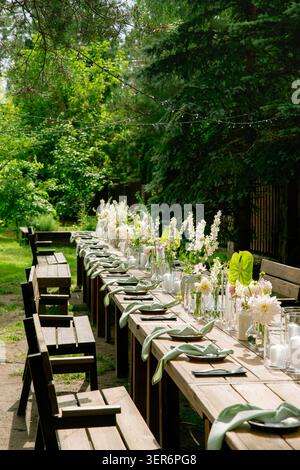 Long outdoor garden wedding table setting with white floral arrangements, sage green napkins and candles amid lush pine trees Stock Photo