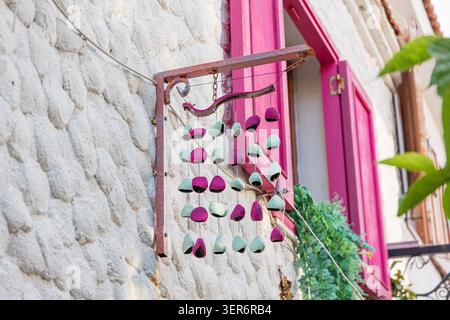 Decorative wind chime with pink and green pieces hanging on a rustic bracket next to vibrant pink shutters on a textured stone wall Stock Photo