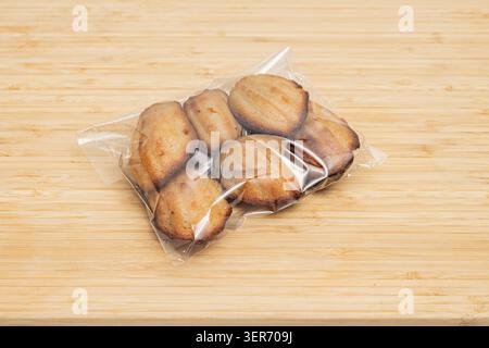 Six golden brown shell-shaped tea cookies sealed in a clear plastic package rest on a light bamboo wooden surface. Stock Photo