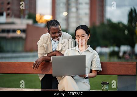 Diverse businesswomen collaborating on laptop outdoors in city park Stock Photo