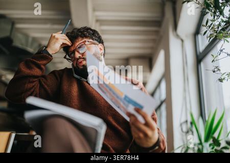 Stressed businessman reviewing documents during a phone call in a modern office workspace Stock Photo