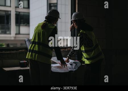 Two female engineers reviewing blueprints at a construction site window wearing safety vests Stock Photo