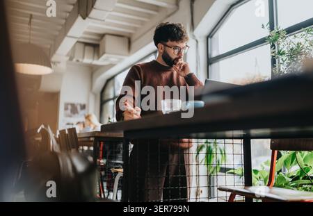 Young businessman working on laptop and taking notes while standing in a modern cafe workspace Stock Photo