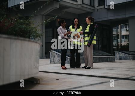 Female architects and engineers in high-visibility vests discussing building plans outside an office complex Stock Photo