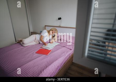 Young woman writing in notebook on bed in cozy bedroom with pink bedding Stock Photo