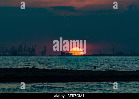 Sun setting behind industrial port cranes and wind turbines in ocean creating energy Stock Photo