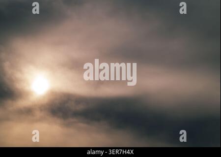 Radiant sun peeking through heavy, dark, and textured grey clouds in the sky. Stock Photo