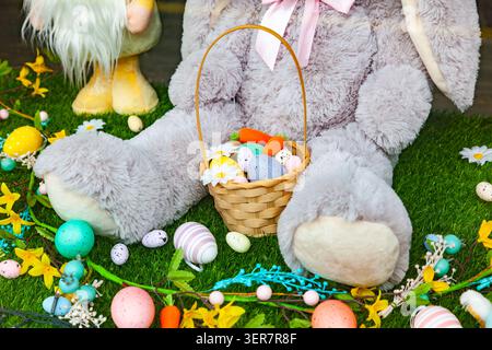 Big plush Easter bunny holding a wicker basket with colorful painted eggs and carrots on green grass Stock Photo