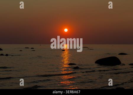 Red sunset over calm sea with a glowing sun reflection on the water and dark coastal rocks along the shoreline Stock Photo