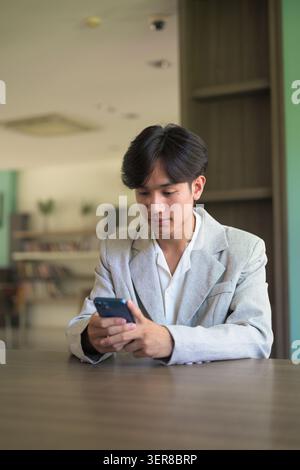Authentic vertical lifestyle portrait of a young non-binary Thai Isan professional or student sitting at a desk using a phone. Concentrating on a smar Stock Photo