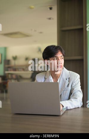 Authentic vertical lifestyle portrait of a young non-binary Thai Isan professional or student working on a computer and looking away thoughtfully. Pau Stock Photo