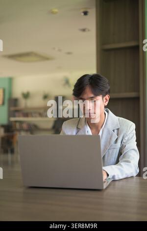 Authentic vertical lifestyle portrait of a young non-binary Thai Isan professional or student typing on a laptop computer. Focused remote work, studyi Stock Photo