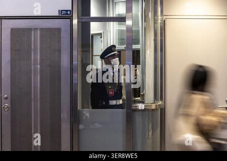 Tokyo, Japan - 25 January 2026: View of a security guard in uniform and face mask standing inside a glass booth while a blurred pedestrian walks past Stock Photo